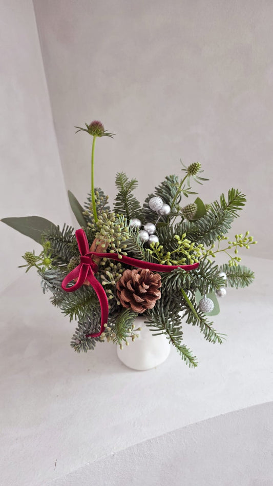 Small Christmas flower arrangement with greenery, pine cones, and a red ribbon on a light gray background.