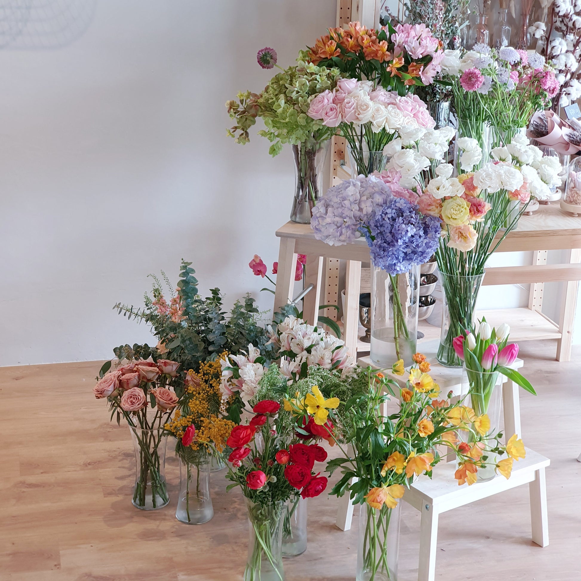 Various flower arrangements in vases on a wooden floor and table.