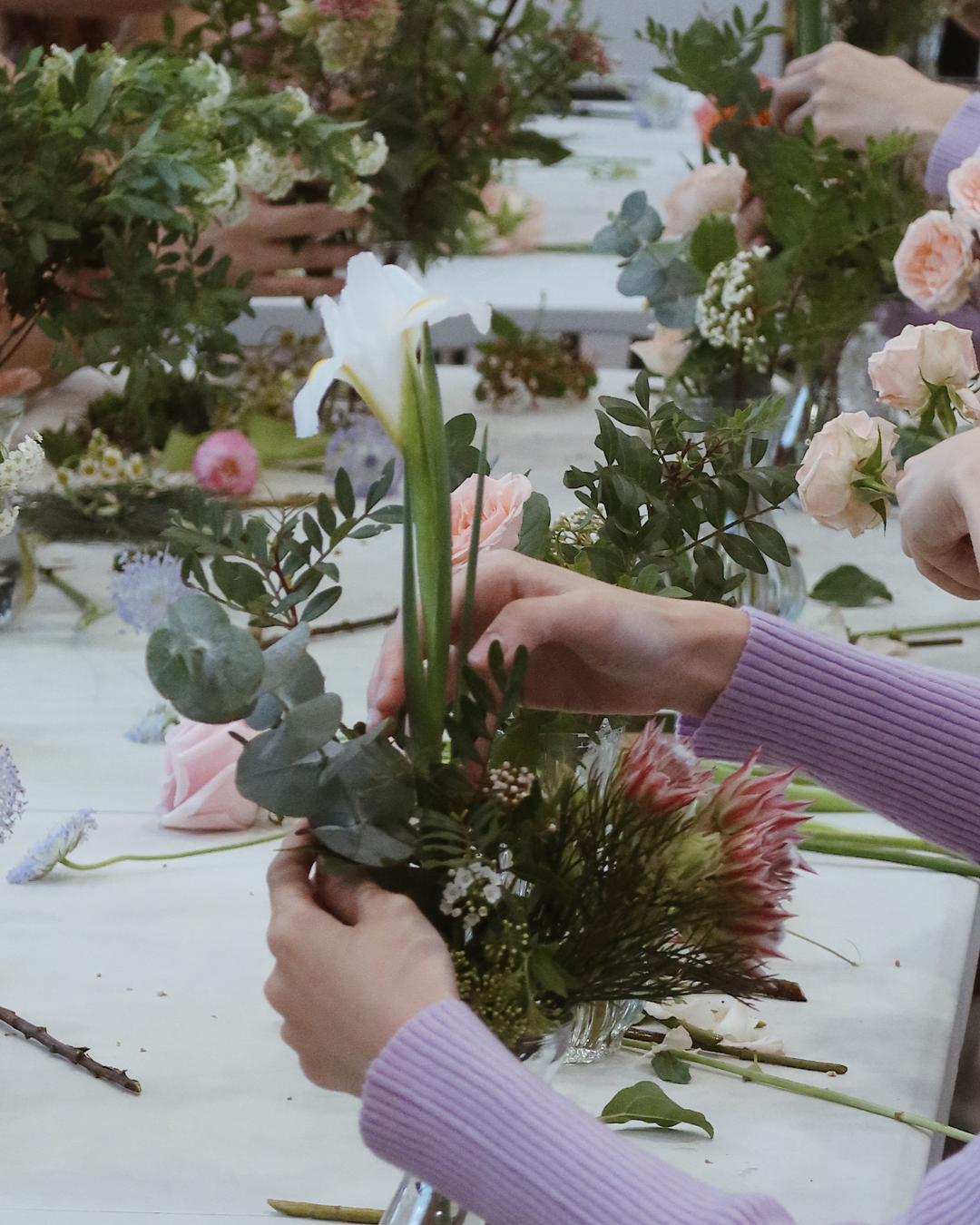 People arranging flowers at a table with a focus on hands and floral materials.