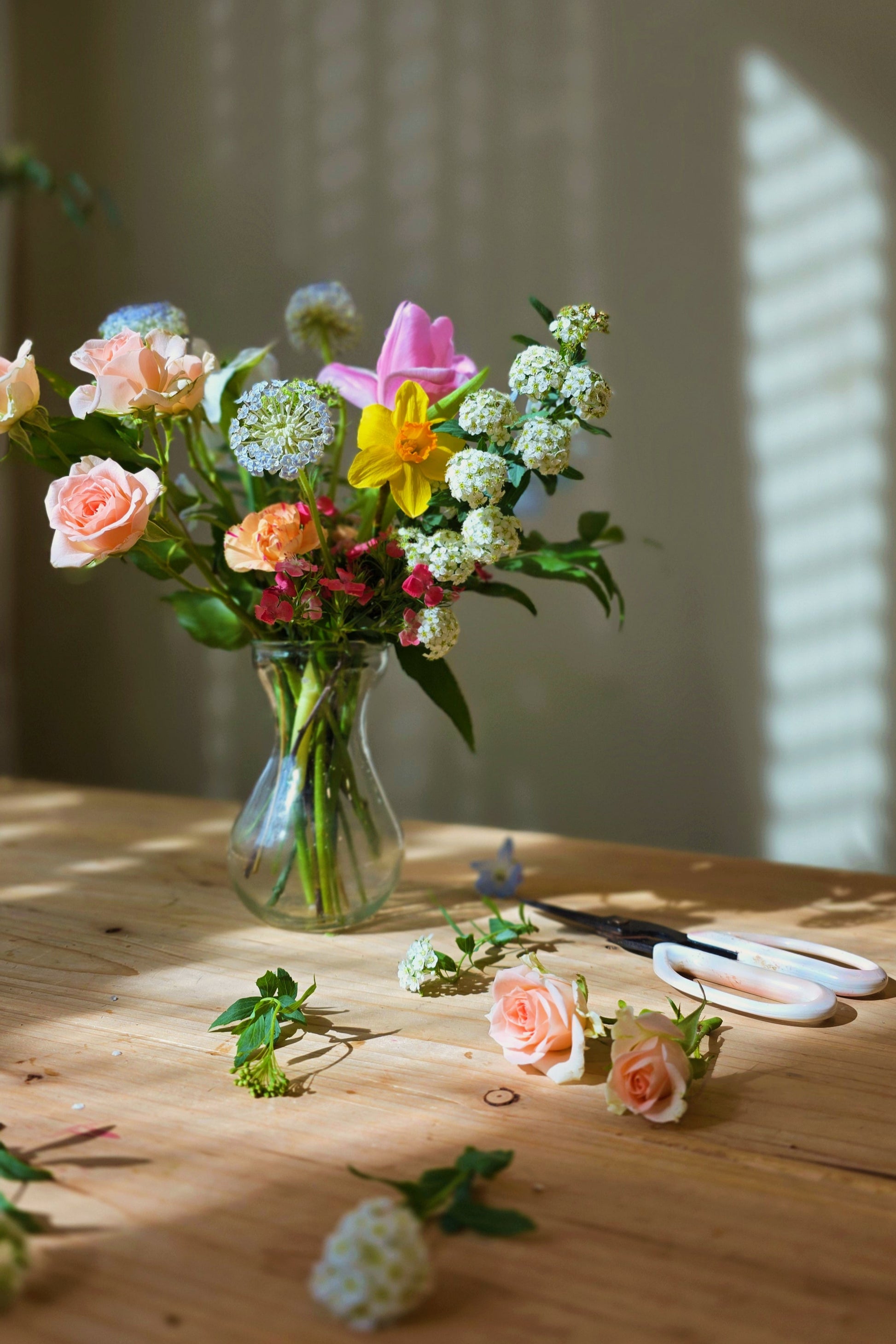 A vase arrangement of fresh flowers including tulips, roses and other foliage, arranged on a wooden table with a window in the background.