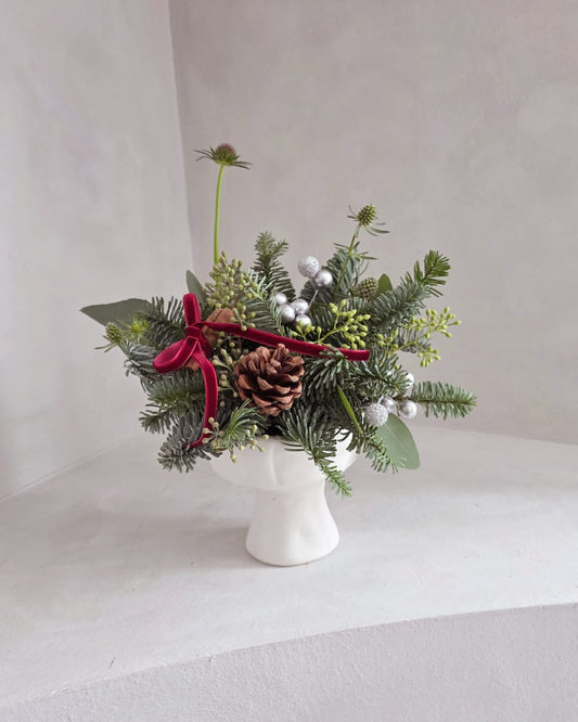 Christmas-themed floral arrangement with greenery, pinecones, and red ribbons in a white vase on a light gray background.