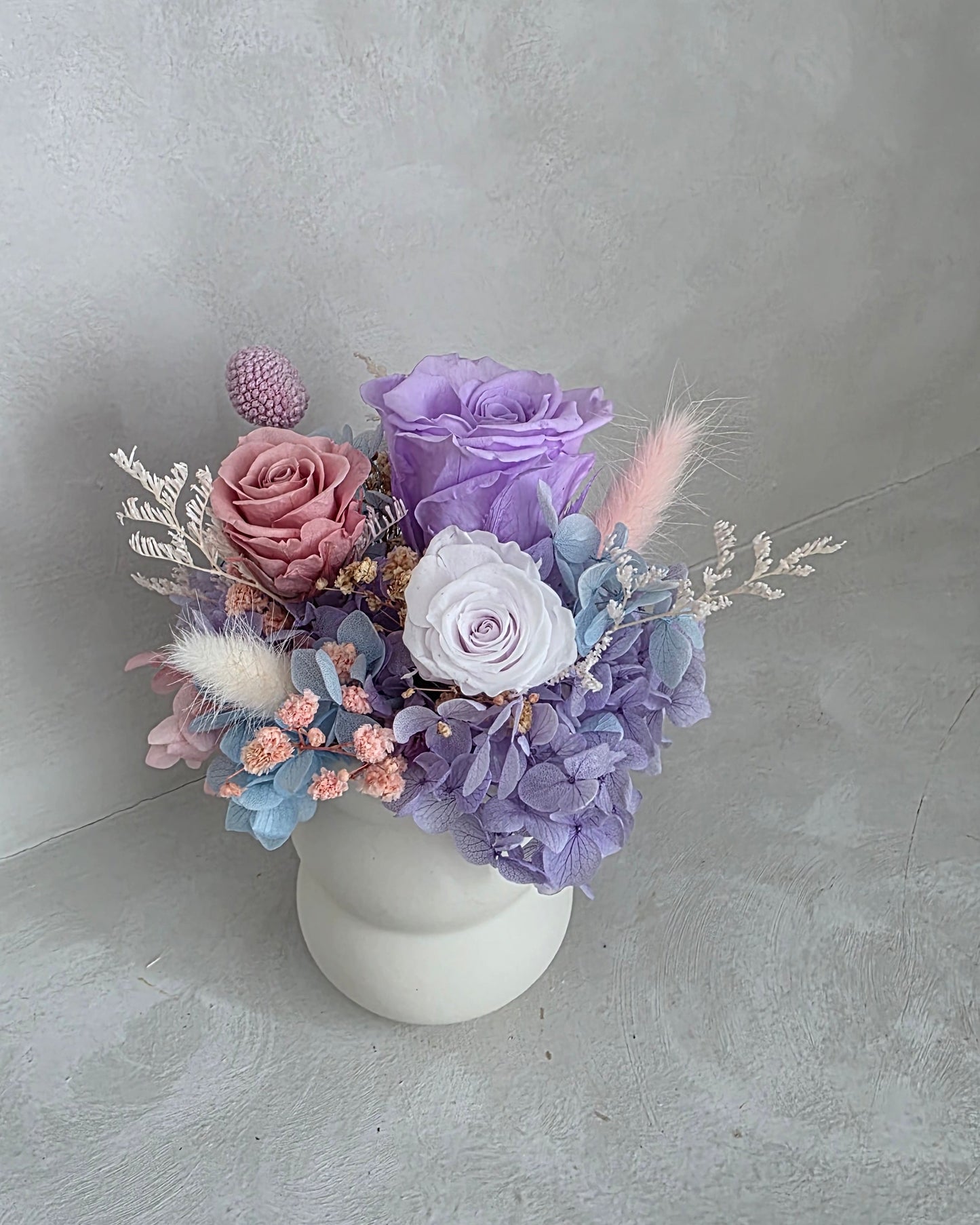 Preserved floral arrangement with purple and pink flowers in a white ceramic vase 