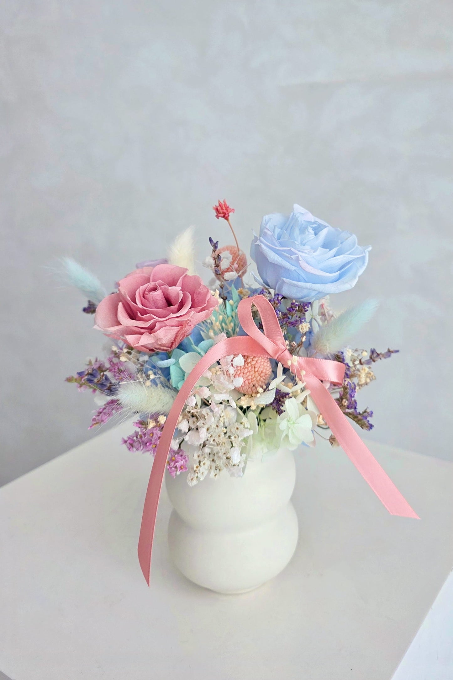 Small arrangement with pink and blue preserved flowers in a white vase on a light gray background